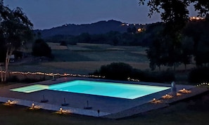 A rectangular swimming pool with a surrounding deck, surrounded by trees and a distant hill.