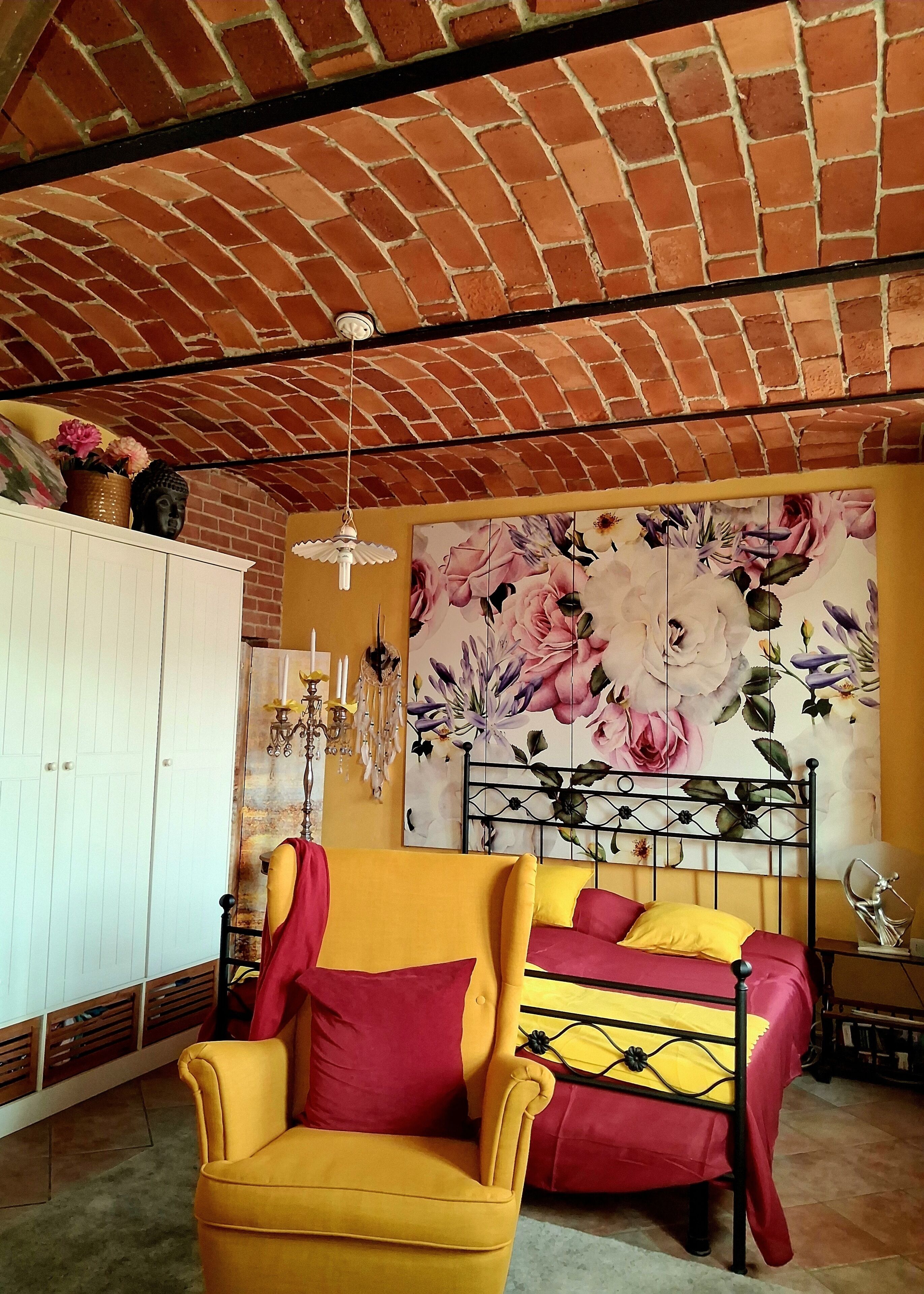 Ein Zimmer mit Backstein-Decke, einem Bett mit Metallgestell, einem gelben Sessel und einer floralen Wandmalerei.