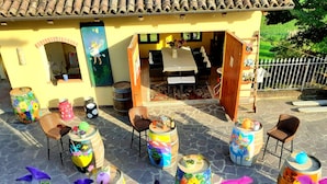 Un edificio amarillo con techo de tejas, mesas y sillas en la terraza, barriles usados como macetas, y un jardín con flores moradas.