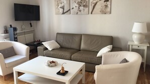 A living room with a beige sofa, white armchairs, a white coffee table, and a flat-screen TV mounted on the wall. - Calle Guerrilleros, entre centro histórico y la zona del Corte Inglès (salamanca)