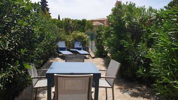 Une terrasse extérieure avec une table et des chaises, entourée d’une végétation luxuriante et dotée d’un sol en pierre.