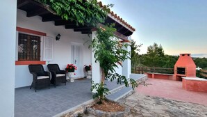 Un edificio blanco con un patio empedrado, dos sillas de mimbre, una planta en maceta y una parrilla.