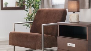 A brown armchair with metal legs, a small wooden side table with a lamp, and a potted plant in a modern room with a window and curtains. - Sunny days in our dreamlike, beautiful Dresden! (Center) (Dresden)
