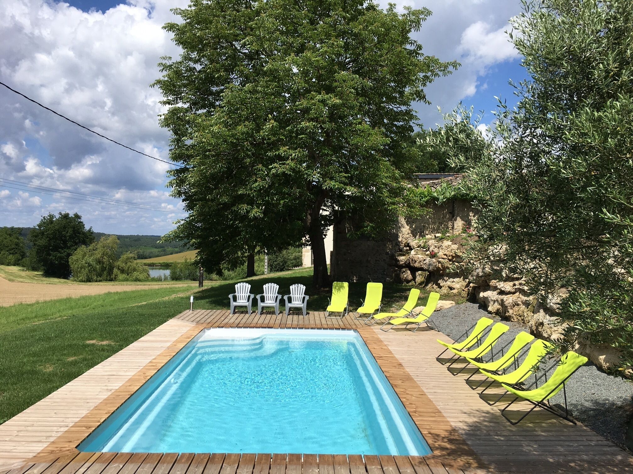 A rectangular swimming pool with a wooden deck, surrounded by yellow lounge chairs and white chairs.