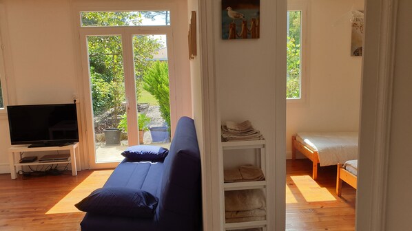 A living room with a blue sofa, a television, a wooden bed, and a view of the outdoors. - QUIET LANDAISE HOUSE ON THE EDGE OF A 3-STAR FOREST (Labenne)