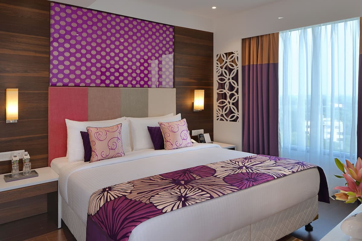 A modern hotel room with a large bed, a nightstand, a wall-mounted lamp, and a view of the city through the window.