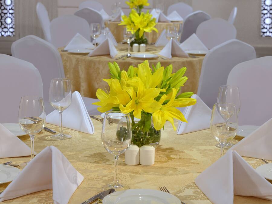 Uma mesa de banquete posta com guardanapos brancos, pratos, talheres e um vaso de flores amarelas.