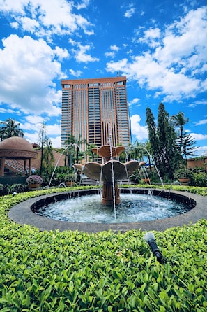 A fountain with water jets in a circular basin, surrounded by lush green foliage. In the background, there is a tall, modern building with a red facade and numerous windows. - KL Service Apartment at Times Square (Kuala Lumpur)