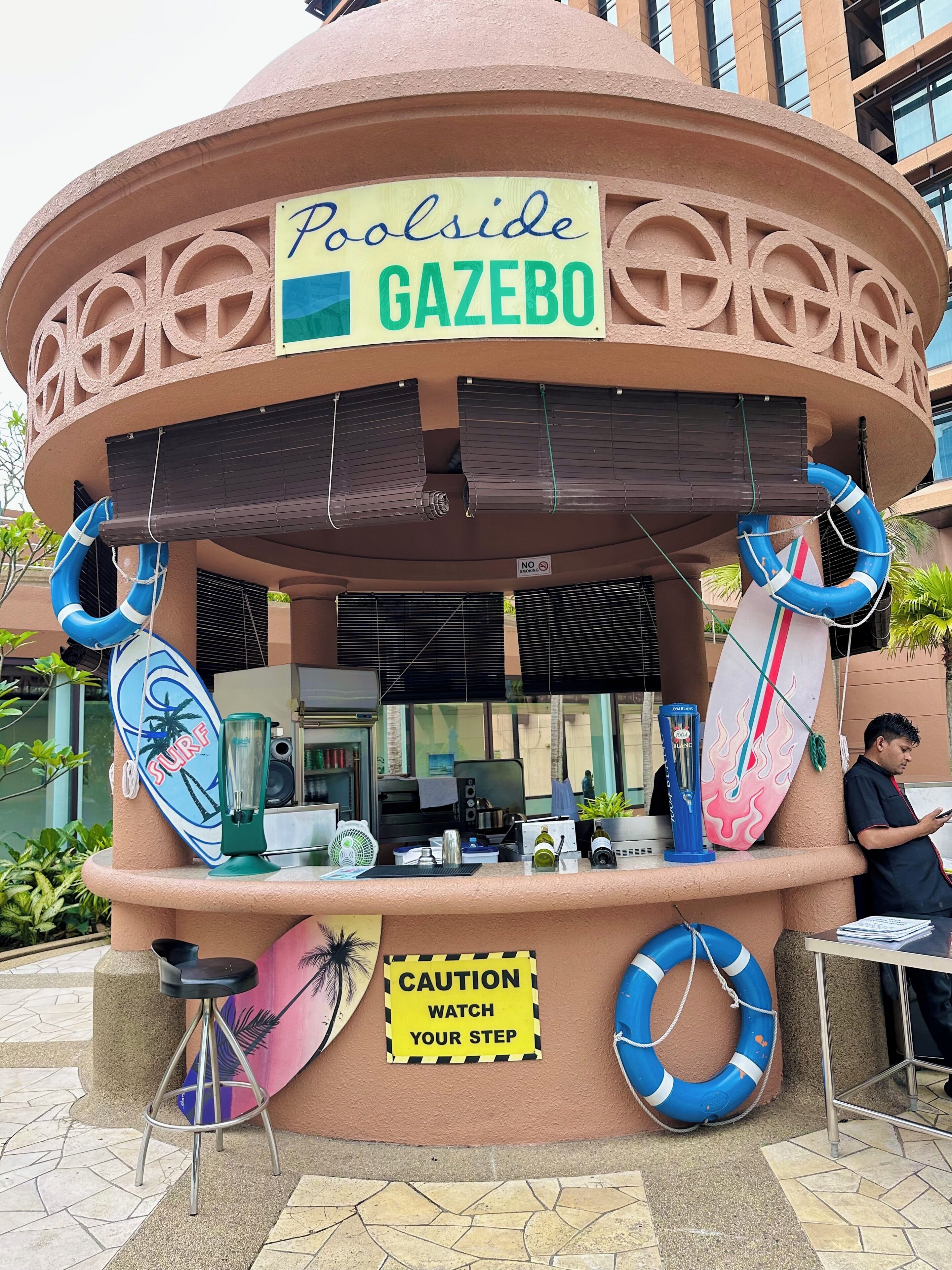 A kiosk named 'Poolside Gazebo' with a caution sign, lifebuoys, and a surf-themed sign.
