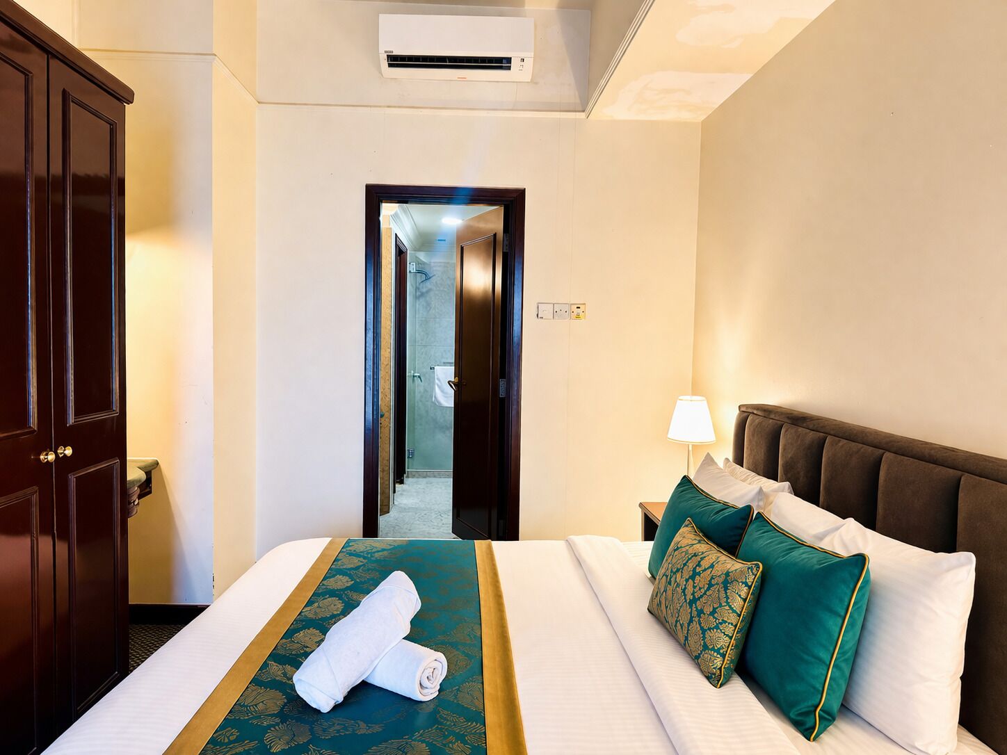 A hotel room with a bed, a nightstand, a lamp, a wardrobe, and an air conditioning unit.