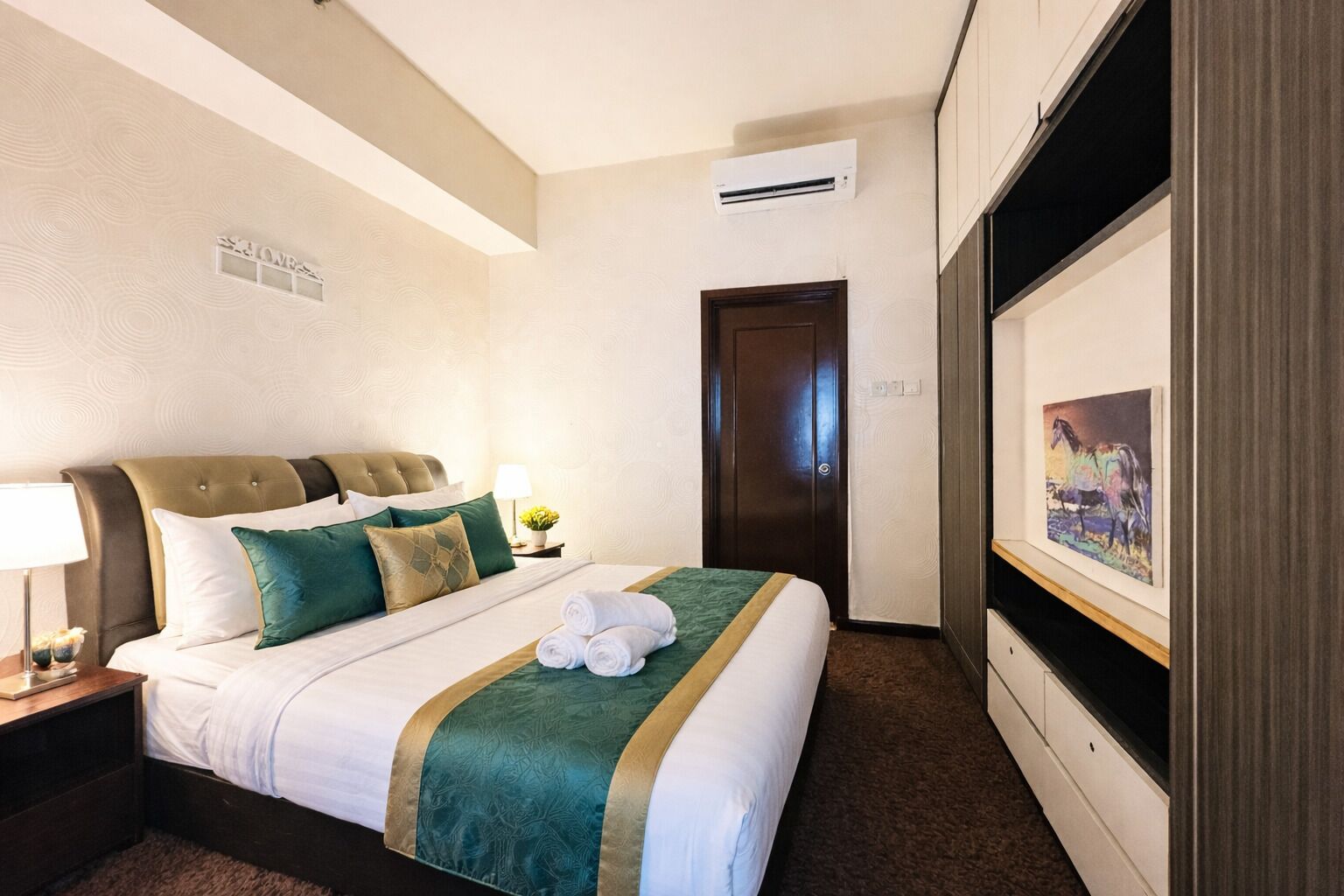 A hotel room with a bed, a nightstand, an air conditioning unit, a door, and a painting on the wall.
