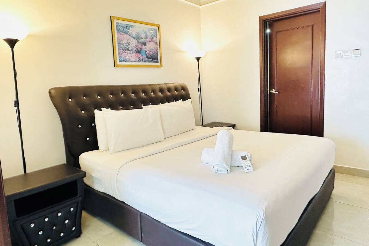 A hotel room with a bed, two bedside tables, a nightstand, a door, and a painting on the wall.