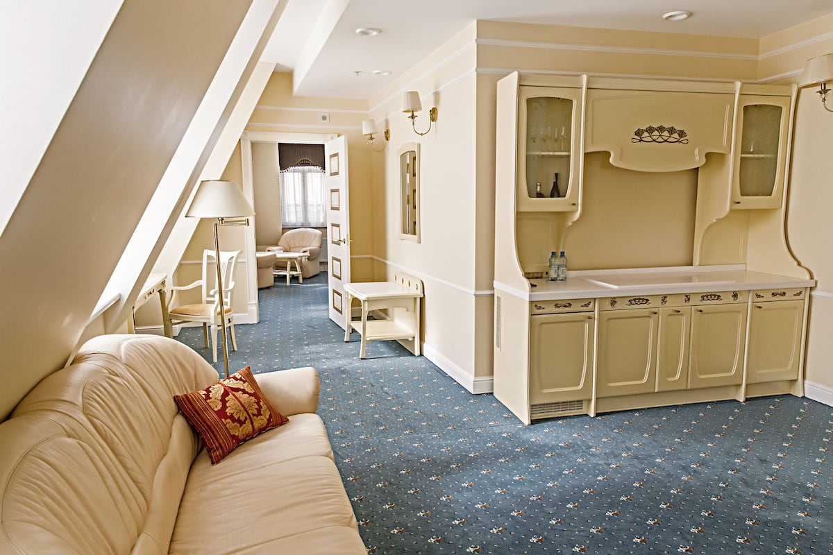 A hotel room with a sofa, a small table, a lamp, a kitchenette, and a bathroom visible through an open door.