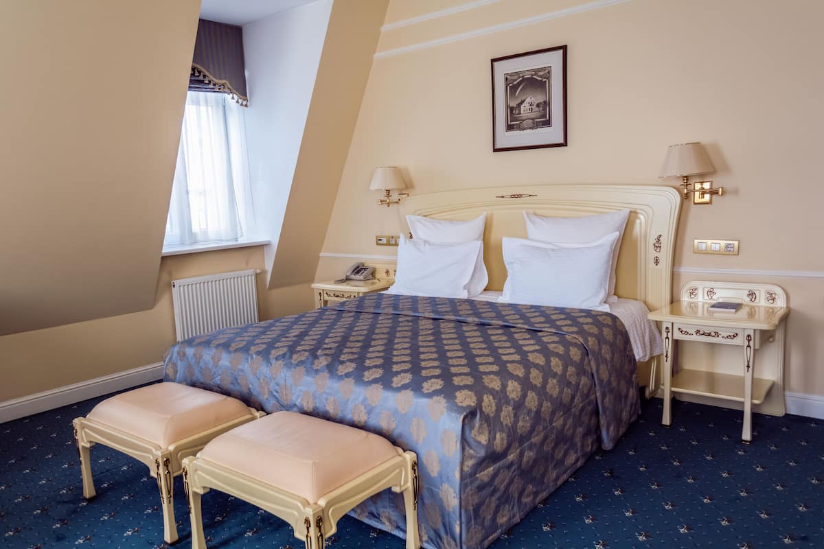 A hotel room with a large bed, two bedside tables, a window with curtains, a radiator, and a small table with a lamp.