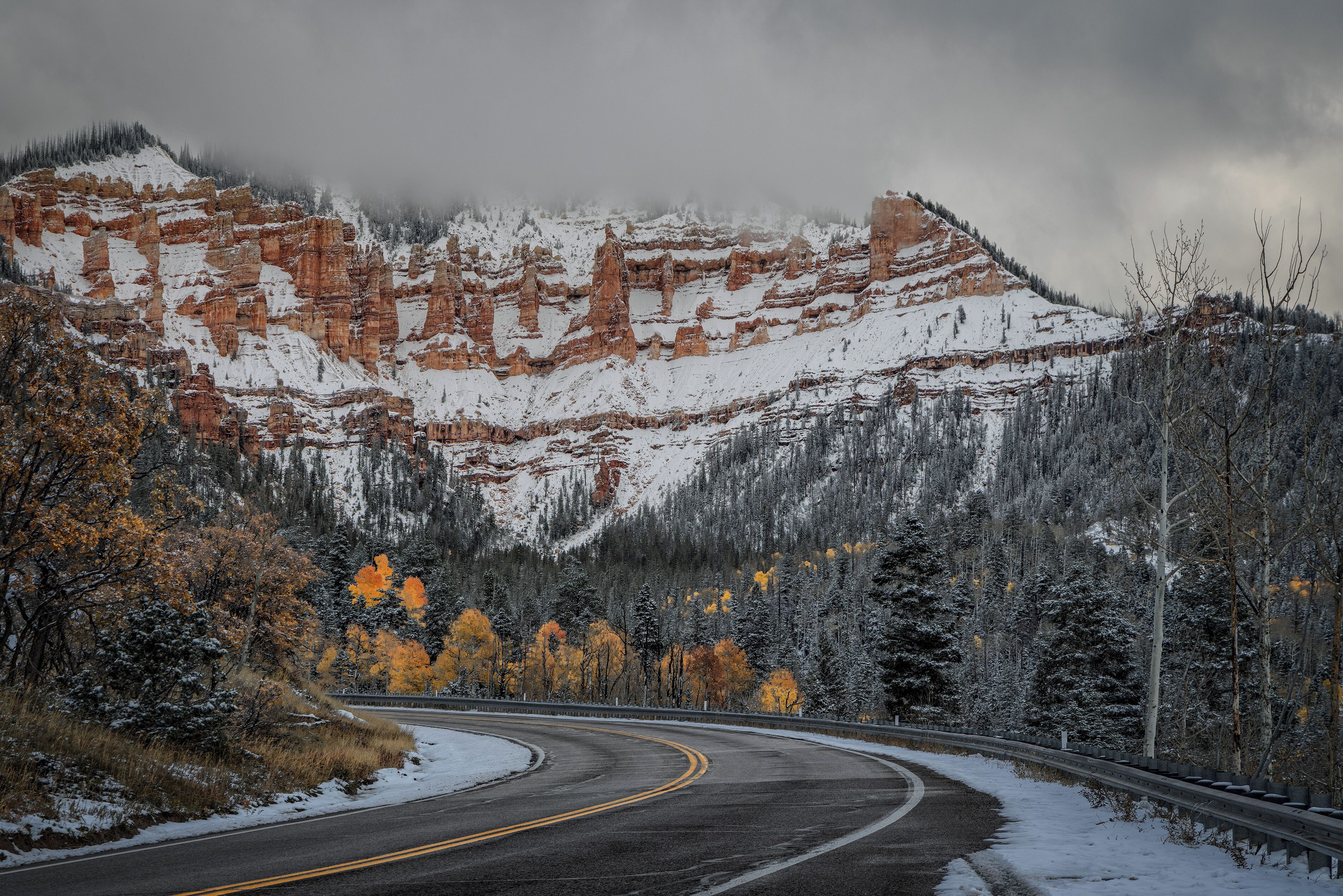 Uma estrada sinuosa serpenteia por uma região montanhosa com neve e árvores de folhagem outonal.