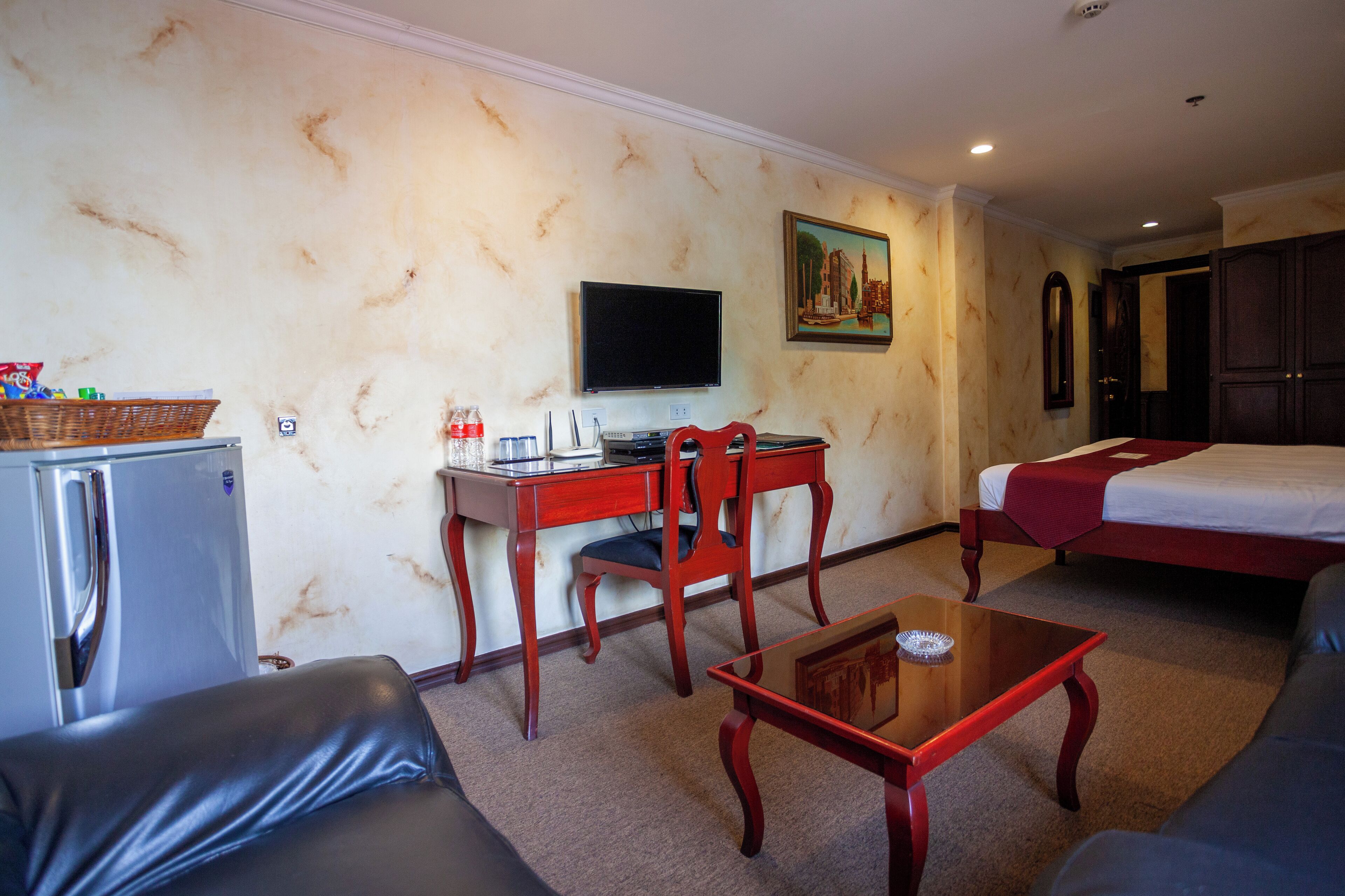 Habitación de hotel con cama, escritorio con computadora, televisor, refrigerador, mesa de centro y sofá.