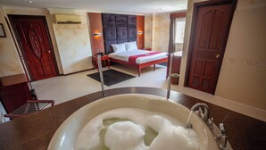 Quarto de hotel com uma banheira grande cheia de espuma, uma cama com cabeceira, uma mesa de cabeceira e uma porta de madeira.