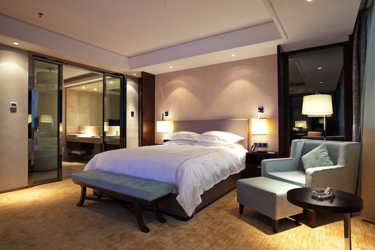 A hotel room with a bed, a bench, a chair, a small table, and a mirror.