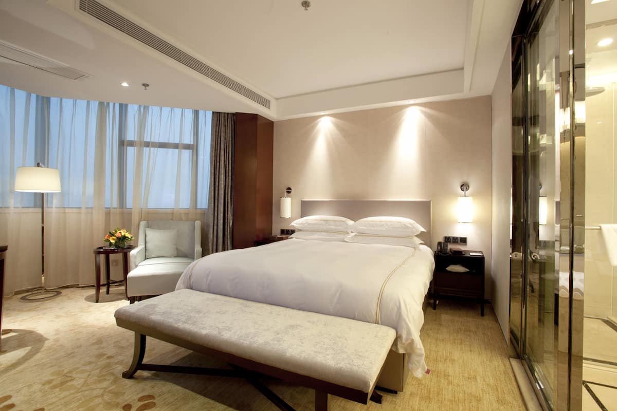 A hotel room with a large bed, a bench, a chair, a bedside table with a lamp, and a glass-enclosed shower area.