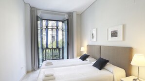 A hotel room with two beds, a large window with a view of buildings, a bedside table with a lamp, and two framed pictures on the wall. - NEW Luxurious Epicenter II Apartment:Free parking, Top location,Free A/C & Wi-Fi (Sevilla)