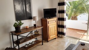 Ein Wohnzimmer mit Fernseher auf einer Holzkommode, einem Blumentopf, einer Lampe und Blick auf Palmen durch ein Fenster.