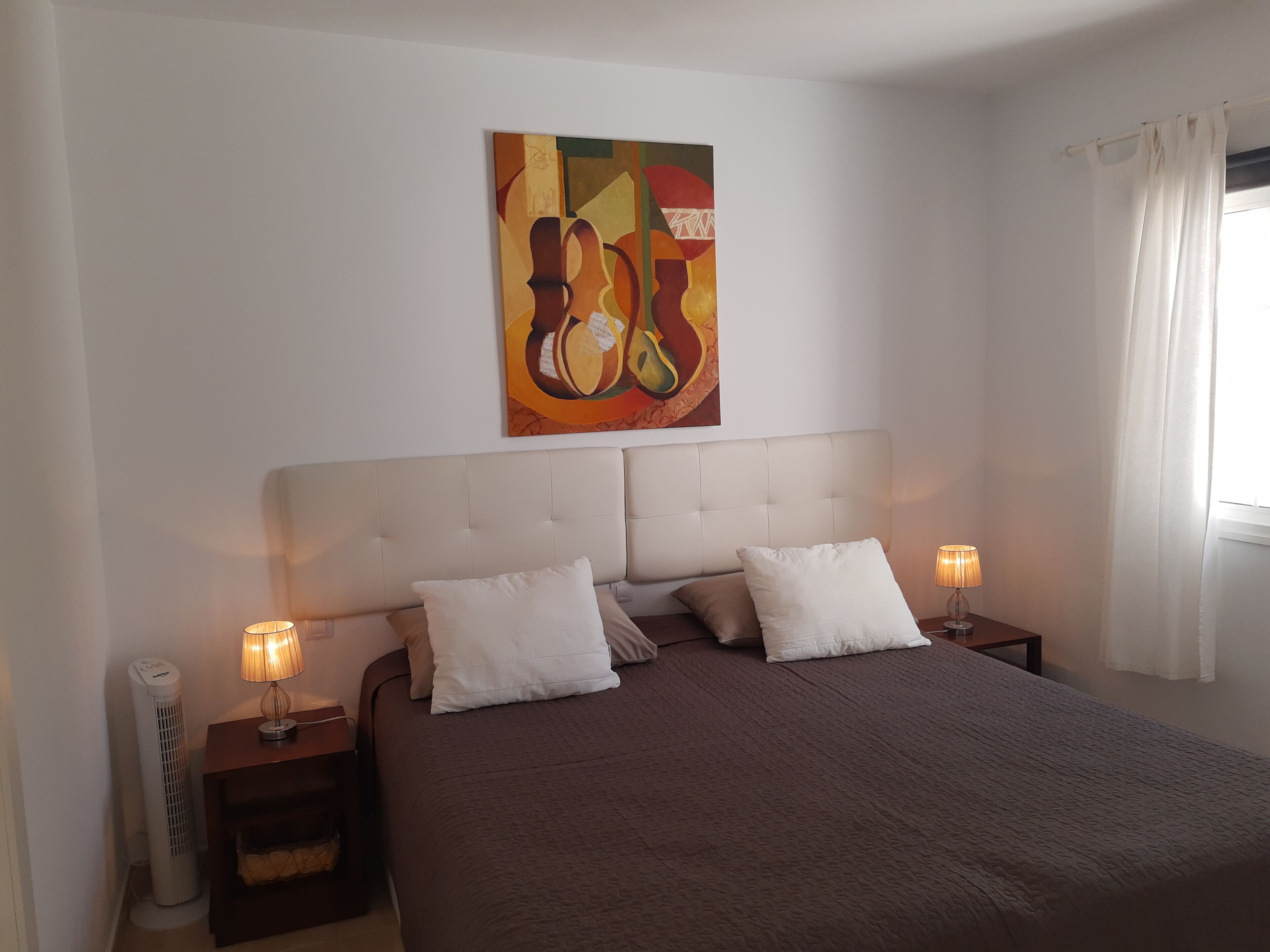 Ein Schlafzimmer mit einem großen Bett, zwei Nachttischen mit Lampen, einem an der Wand hängenden Kunstwerk und einem Fenster mit Vorhängen.