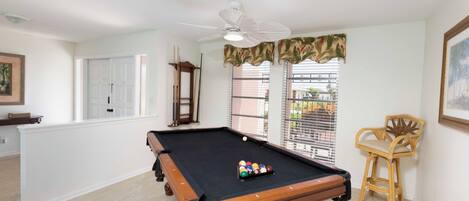 A pool table in a room with a ceiling fan, a window with blinds, and a chair.