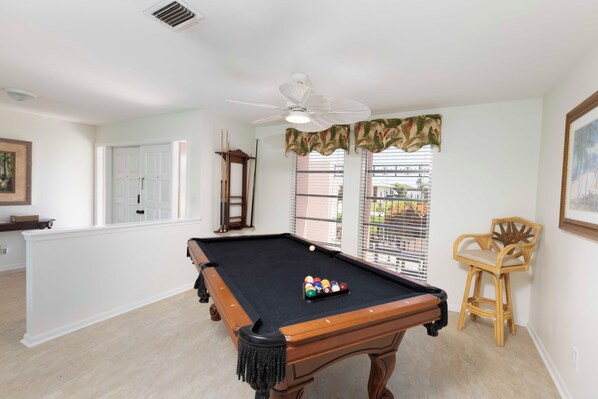 Una mesa de billar en una habitación con ventilador de techo, una ventana con persianas y una silla.