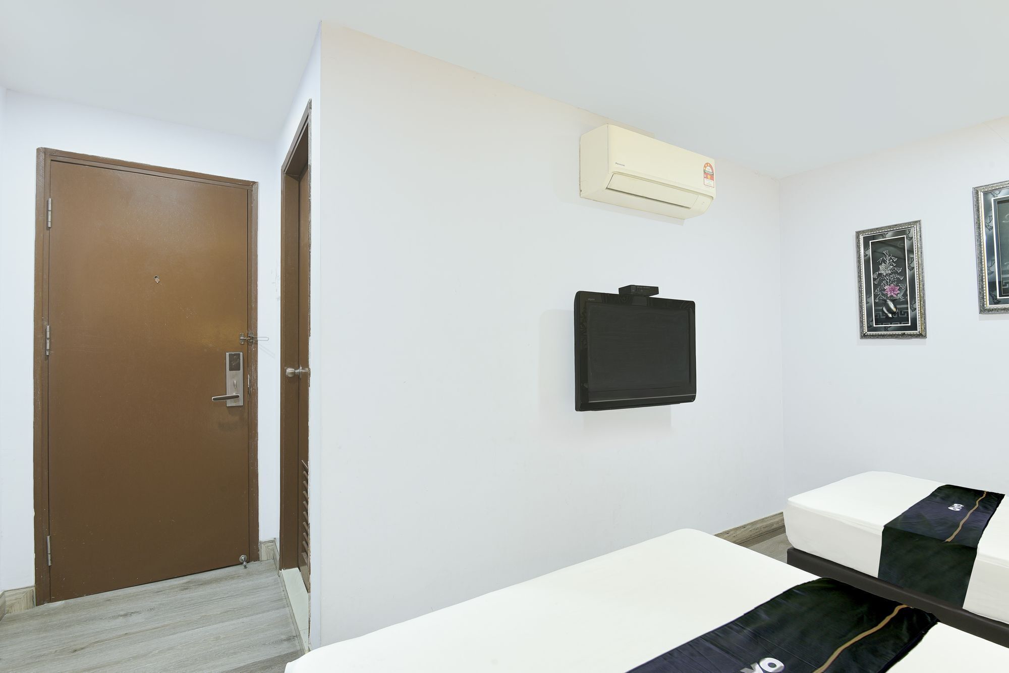 A hotel room with a brown door, a white wall, an air conditioning unit, a black electronic device, and two framed pictures.