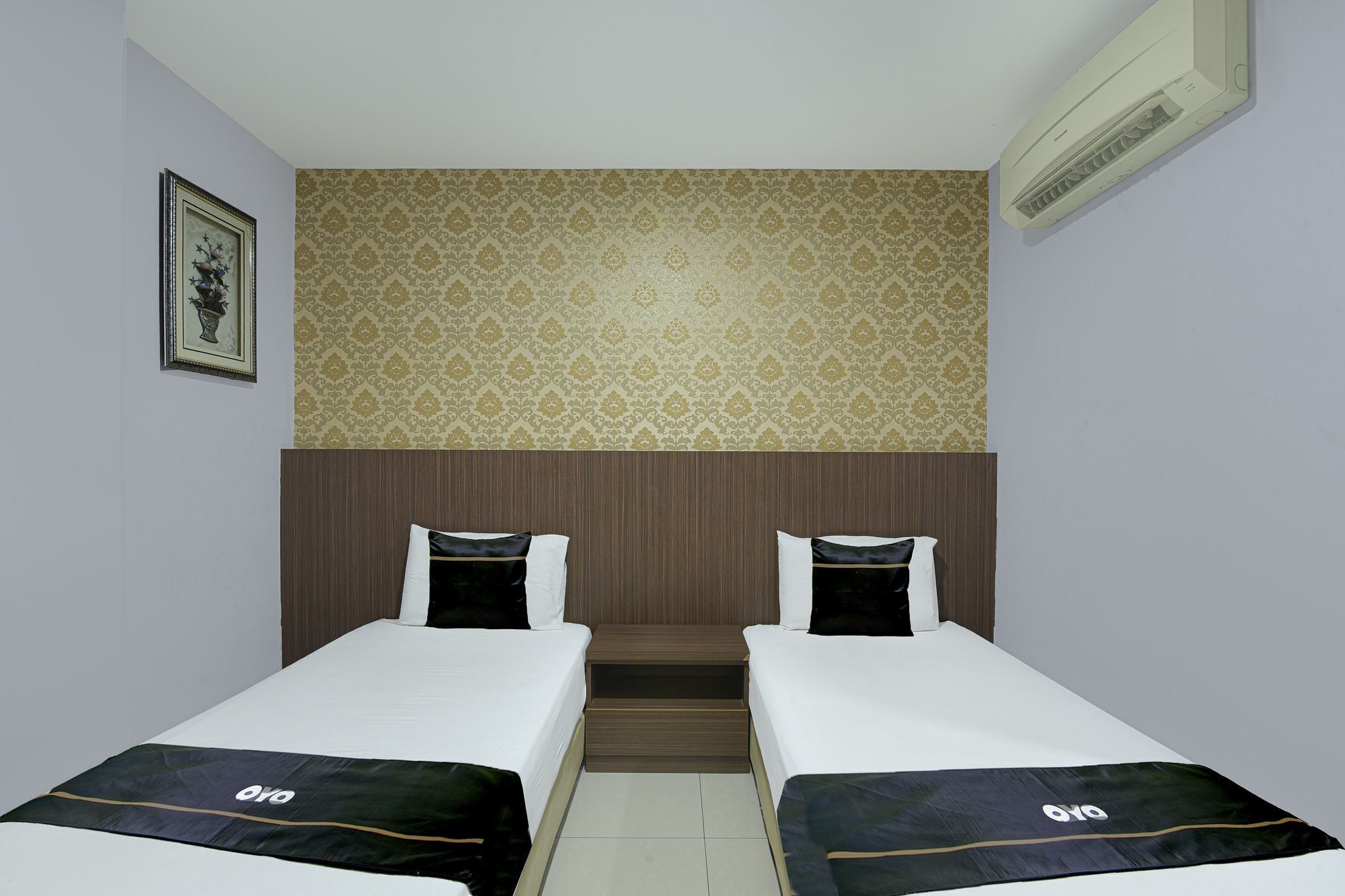 Een hotelkamer met twee eenpersoonsbedden, behang met een patroon, een ingelijst schilderij aan de muur en een ingebouwde airconditioning.
