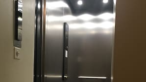 An elevator interior with metallic walls, a control panel, and a glass door. - Piazza Collegiata Bellinzona (Bellinzona)