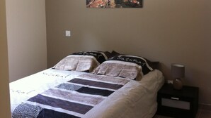 Een slaapkamer met een bed, kussens, een nachtkastje, een lamp en een schilderij van een kustplaatsje.