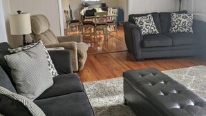 Una sala de estar con un sofá oscuro, un reposapiés y almohadones estampados. Se ve un comedor con mesa y sillas a través de un umbral abierto.