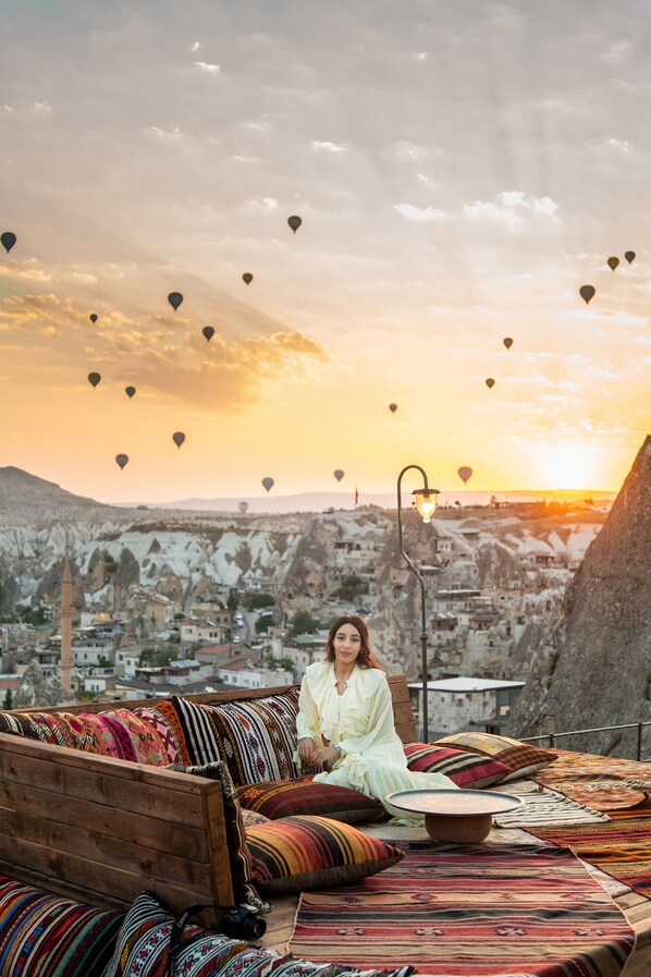 A person sitting on a sofa with colorful cushions, overlooking a cityscape with buildings and a sunset. - Charming Cave Hotel (Nevsehir)