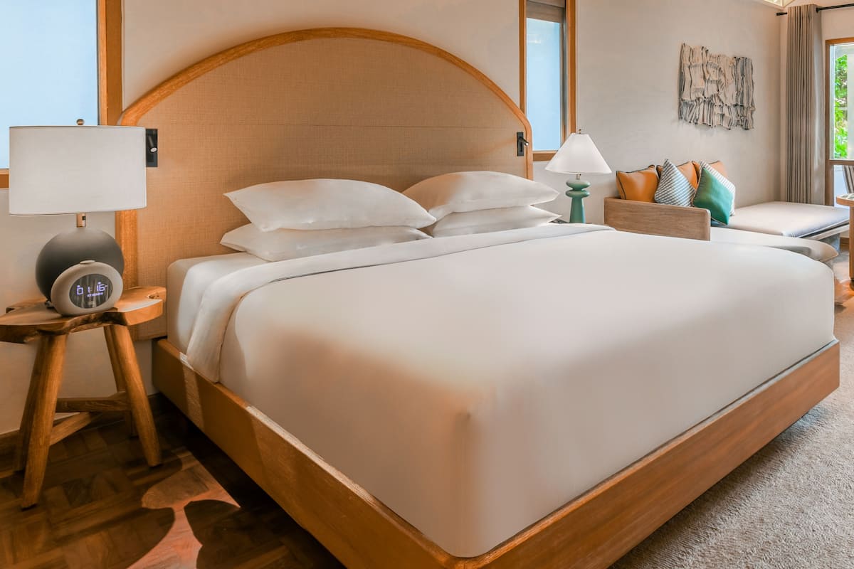 A modern hotel room with a large bed, a bedside table with a lamp, a clock, and a small seating area with cushions.