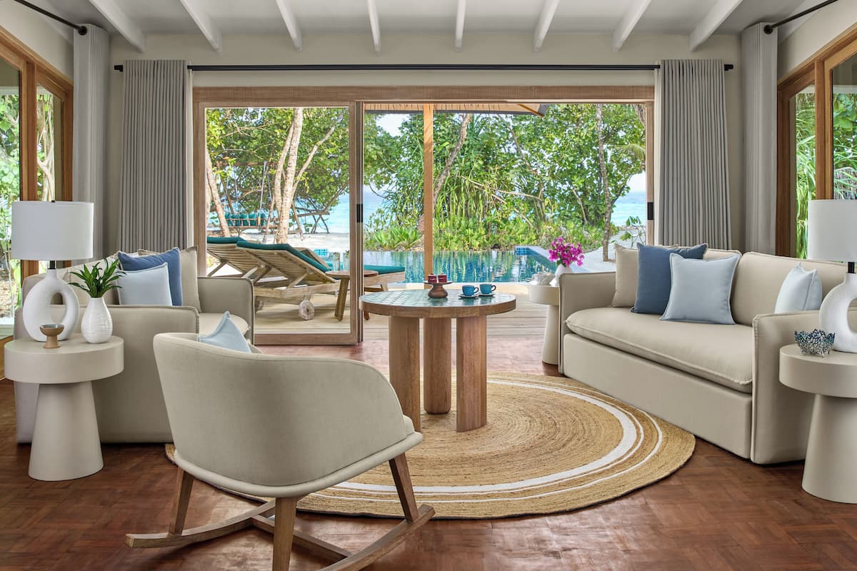 A modern living room with a view of a pool and beach, featuring a sofa, armchair, coffee table, and side tables.