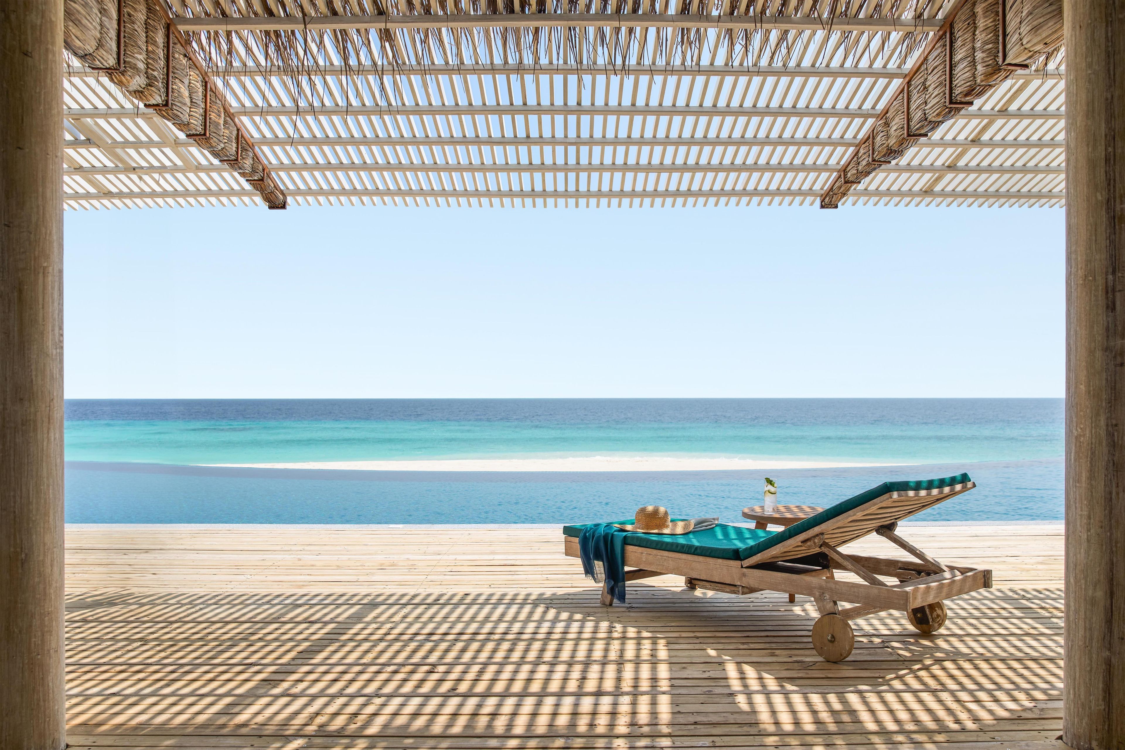 Een houten terras met een ligstoel en een tafel, met uitzicht op een strand met helderblauw water.
