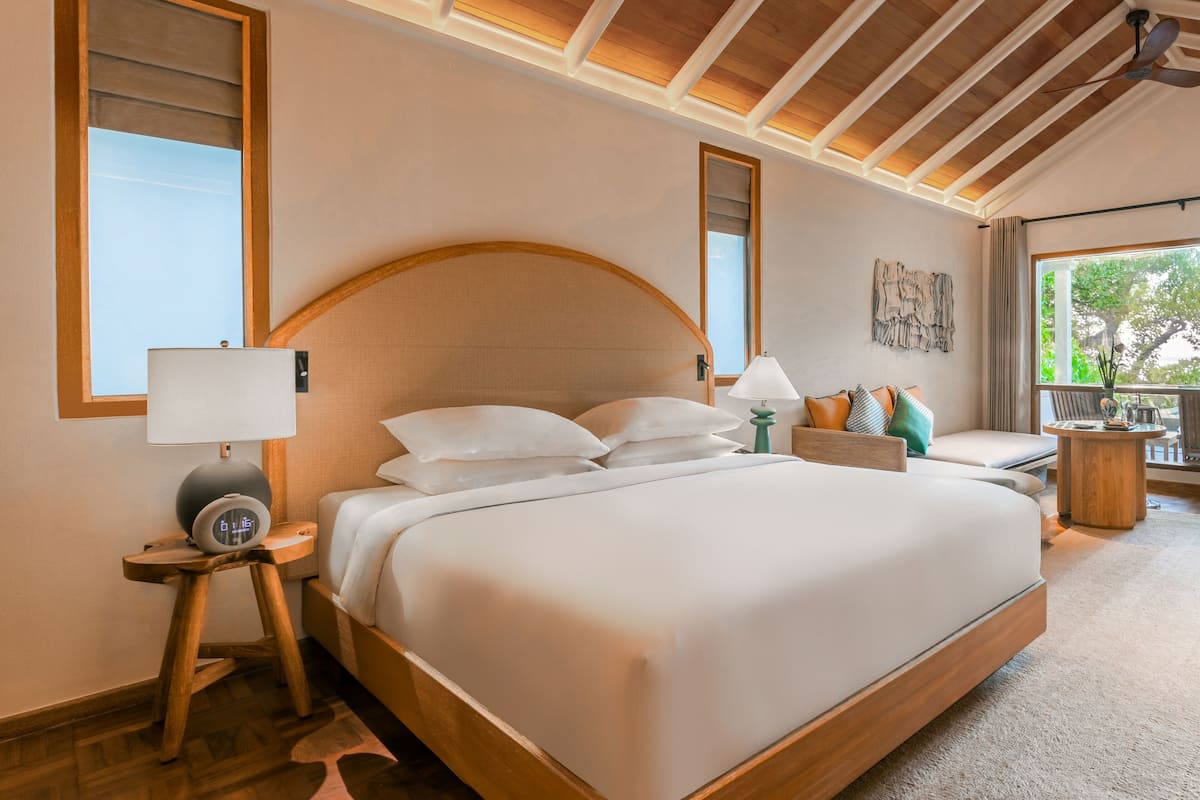 A spacious bedroom with a large bed, a bedside table with a lamp, a small stool, and a view of the outdoors through a large window.