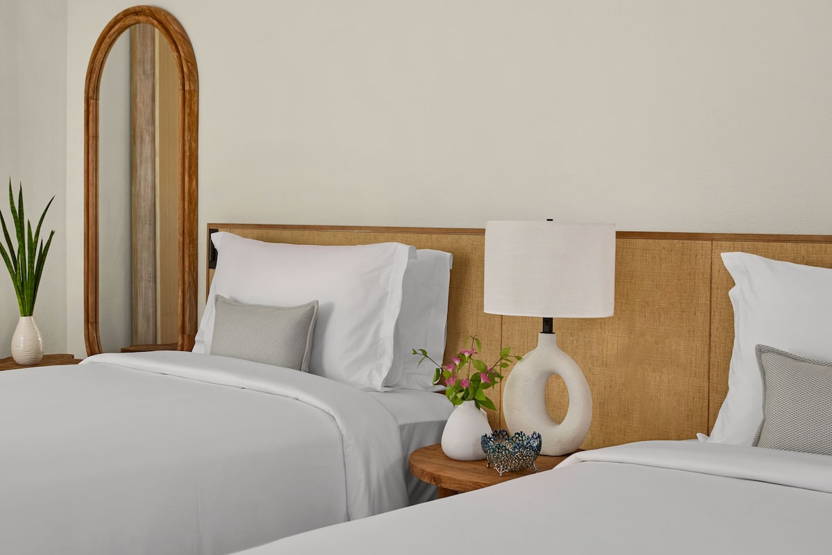 A hotel room with two beds, a wooden headboard, a bedside table with a lamp, a vase with flowers, and a mirror.
