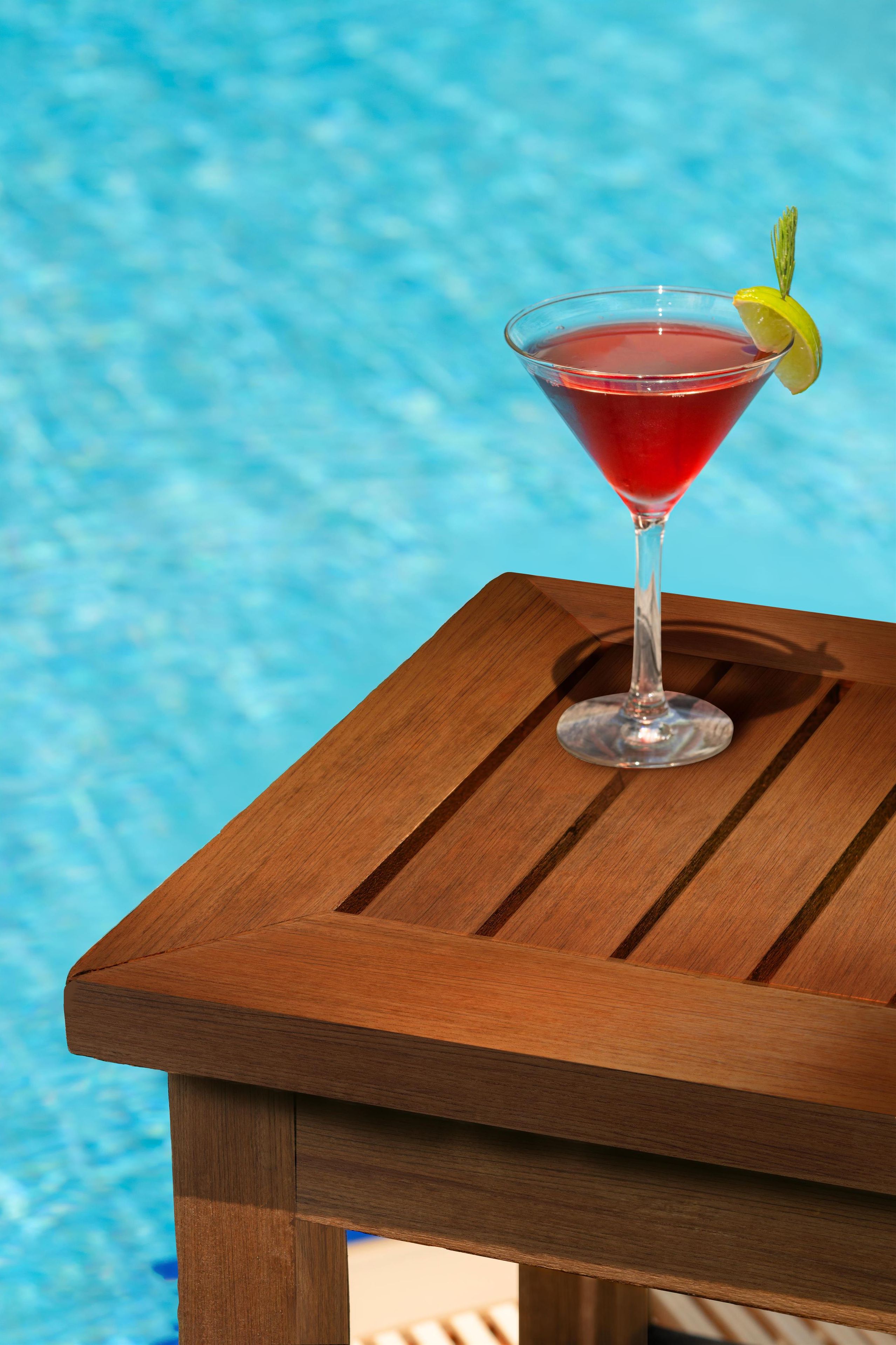 Una copa de martini con una bebida roja y un adorno de limón sobre una mesa de madera junto a una piscina.