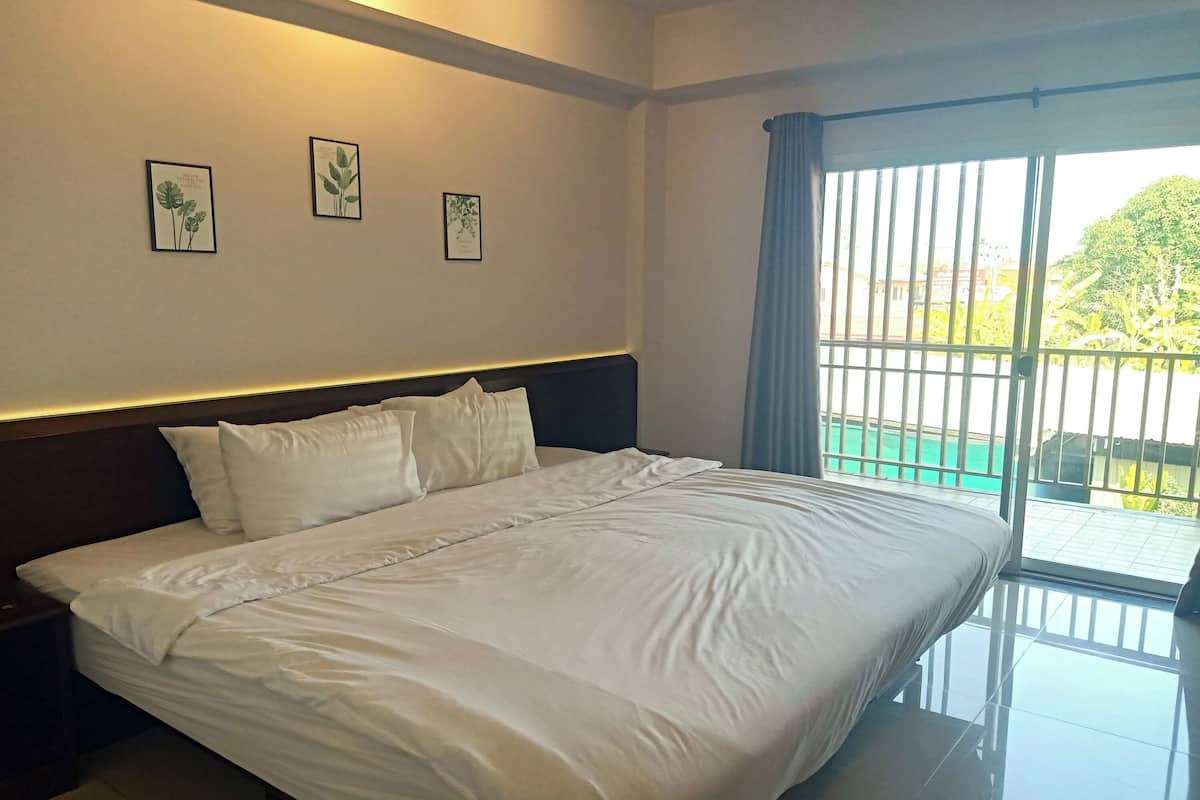 A hotel room with a large bed, a nightstand, and a window with a view of a swimming pool and greenery.