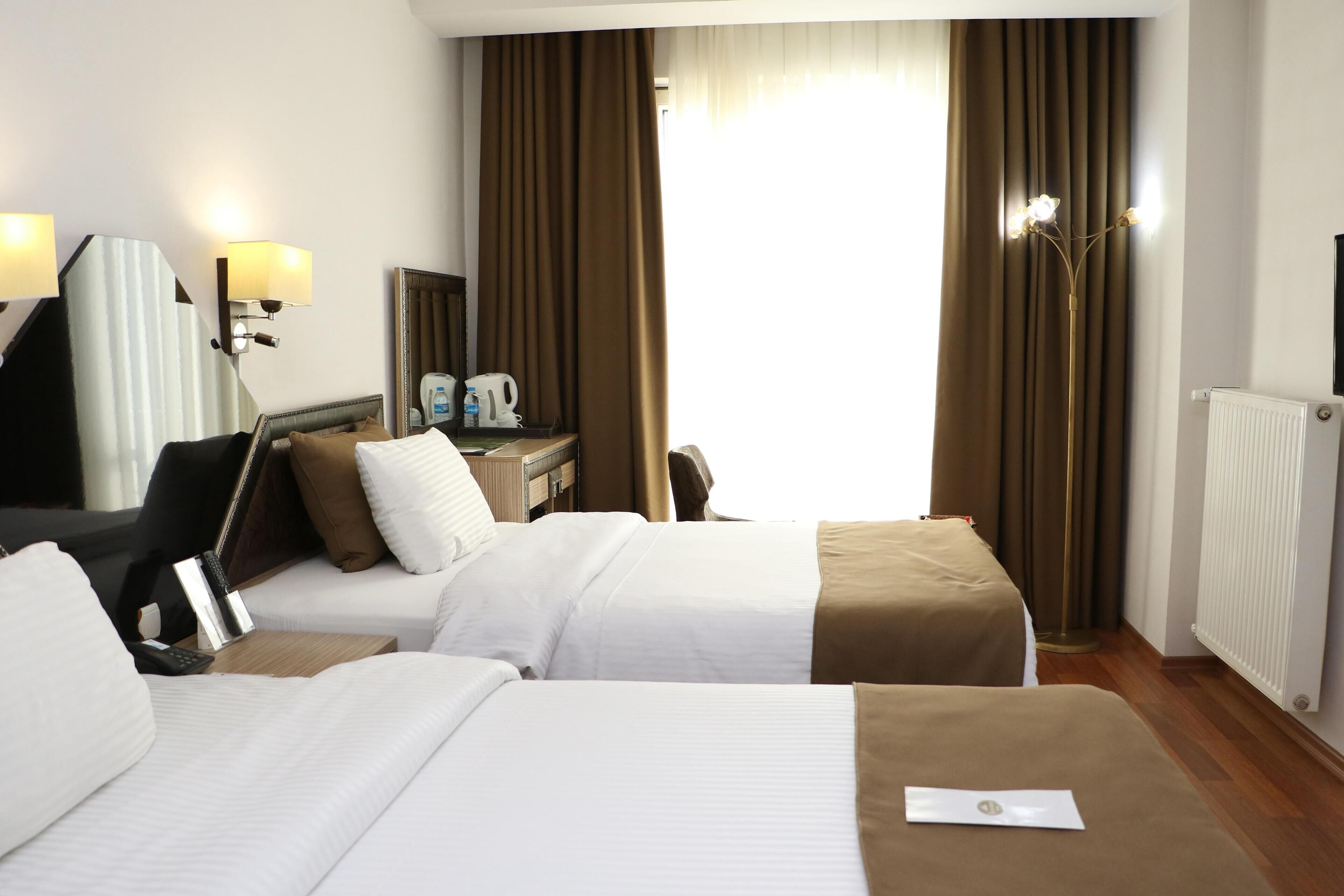 A hotel room with two beds, a desk with a lamp, a mirror, a radiator, and a window with curtains.