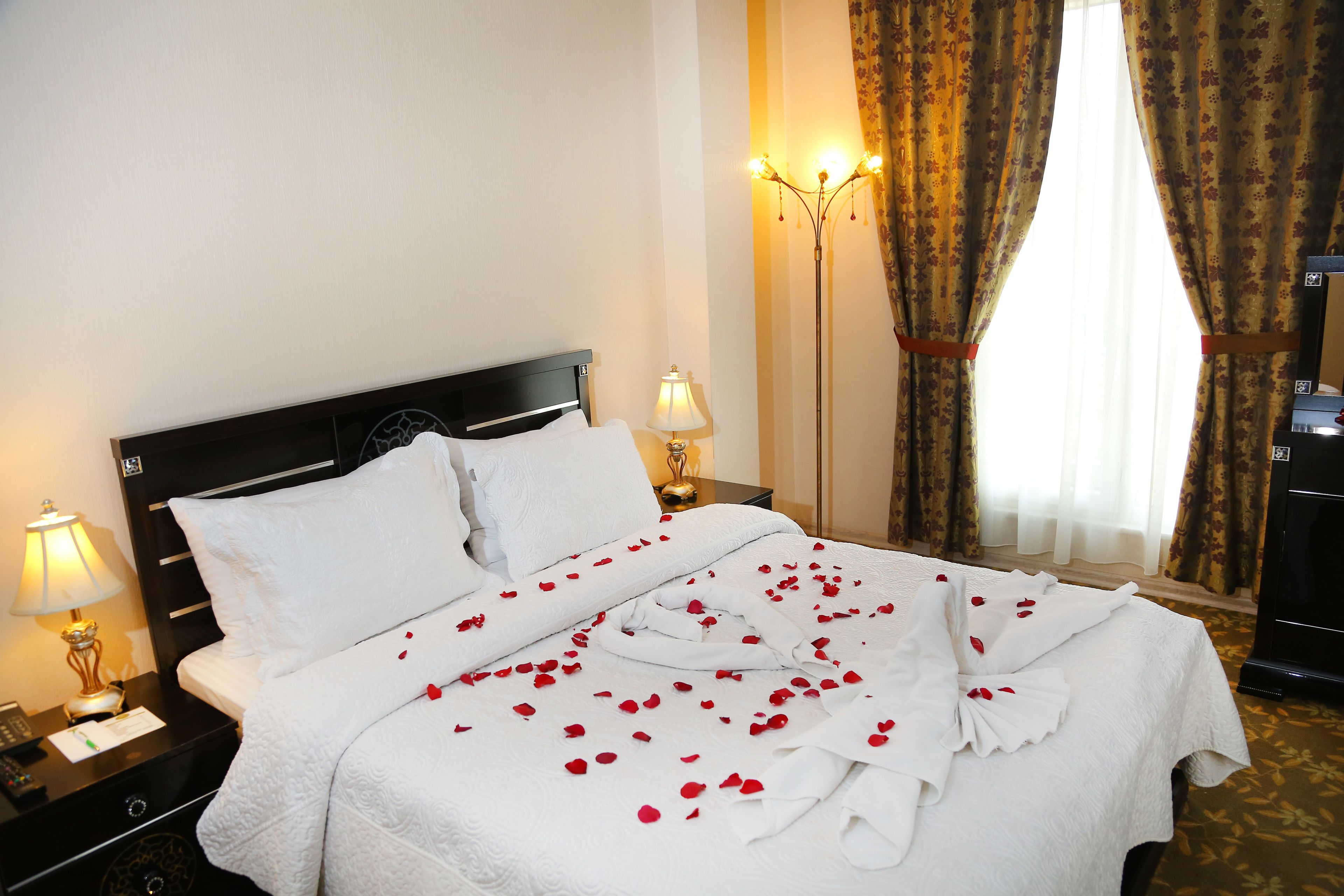 A hotel room with a neatly made bed, a nightstand with a lamp, and a large window with curtains.