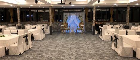 A banquet hall with rows of tables and chairs, a stage with a backdrop, and a ceiling with recessed lighting.