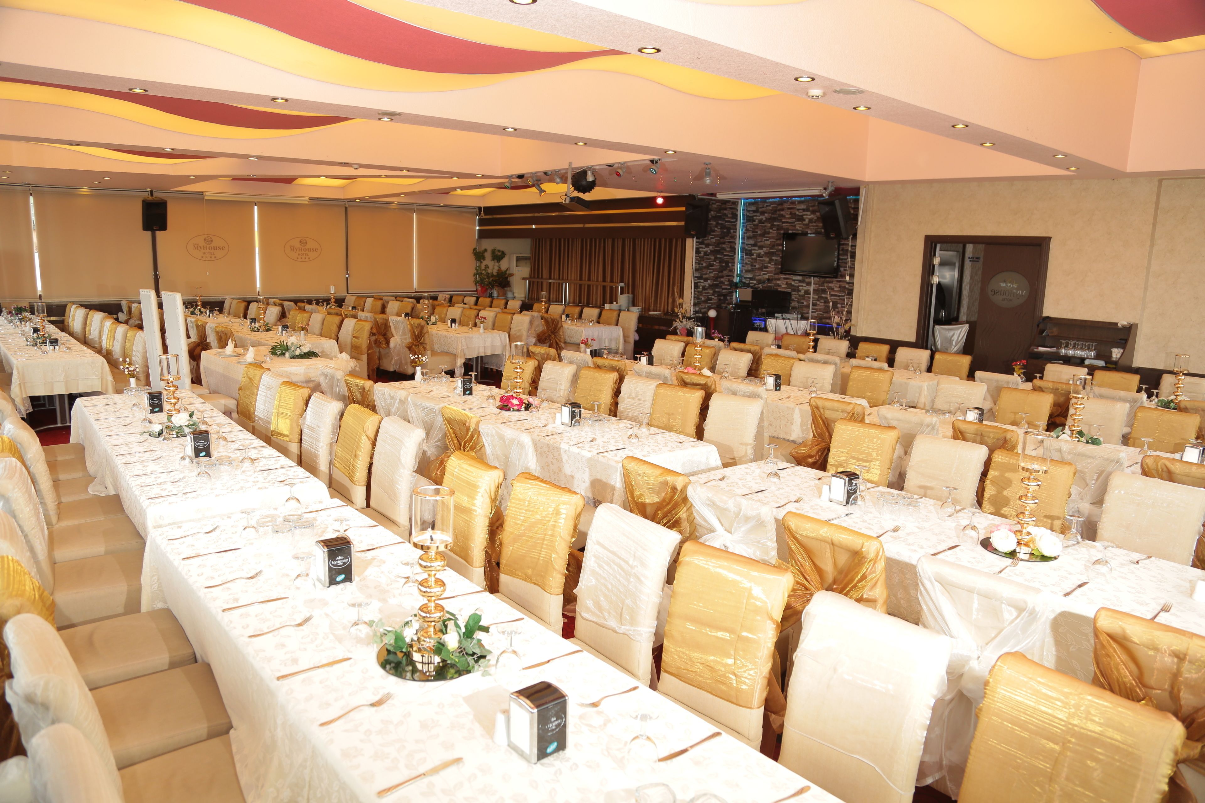 Un salón de banquetes con filas de mesas dispuestas para un evento formal, destacando fundas doradas para las sillas y manteles blancos.
