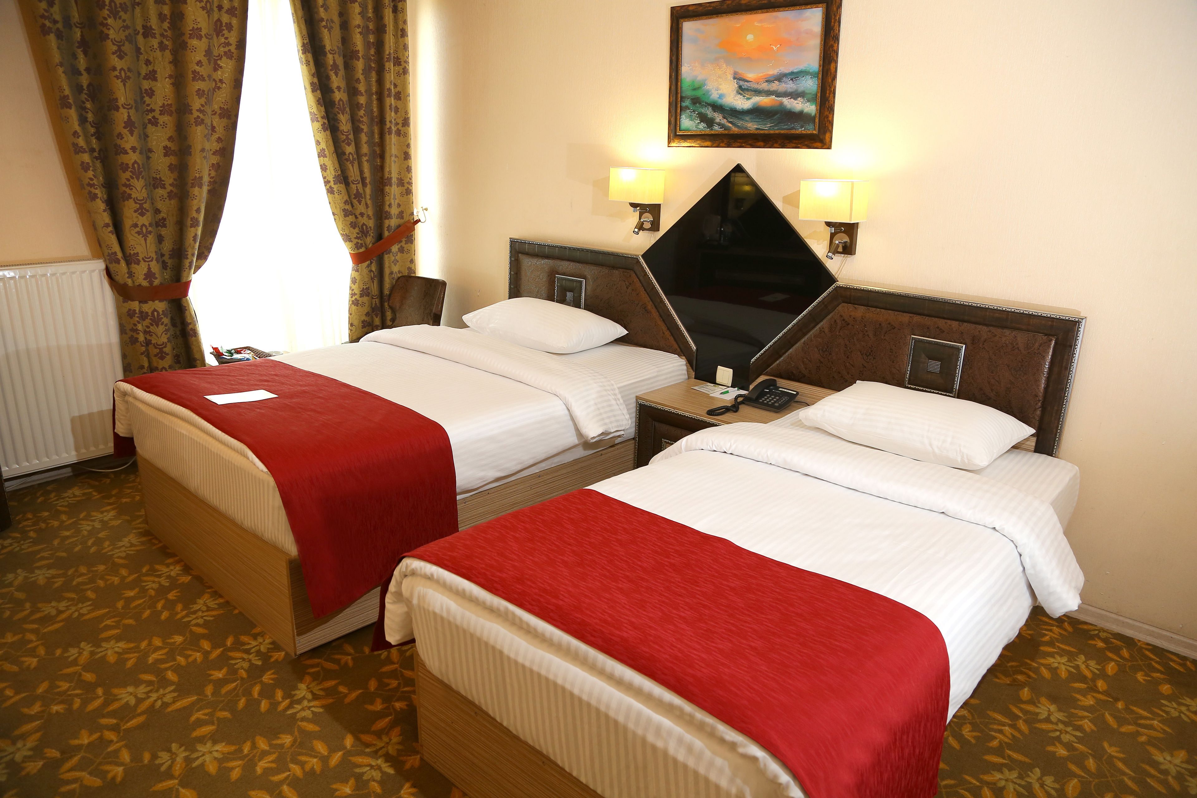 A hotel room with two beds, each with a red blanket, a nightstand with a lamp, and a painting on the wall.