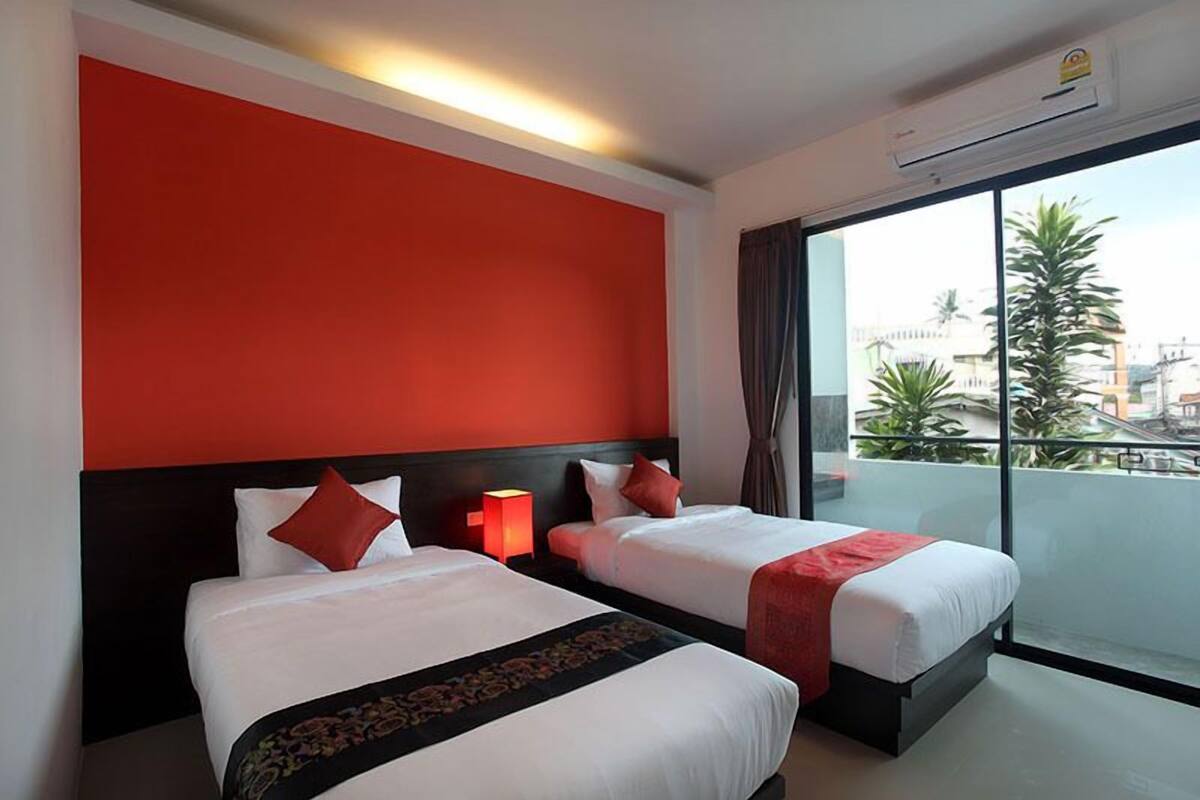 Habitación de hotel con dos camas, una pared de acento roja, un balcón con vistas y un equipo de aire acondicionado.