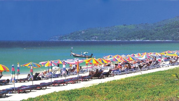 A beach with rows of sun loungers and colorful umbrellas, a boat in the water, and a hilly landscape in the background. - Karon Sino House (Karon)