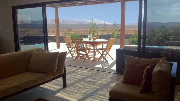 Un salon moderne doté d’une grande porte vitrée donnant sur un patio extérieur, avec un ensemble de mobilier de jardin et une vue sur un paysage montagneux.