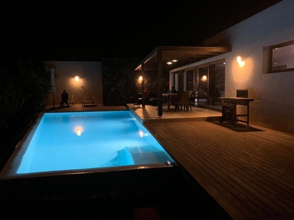 Relax by the pool under the warm glow of wall-mounted lights, surrounded by a comfortable seating area with tables and chairs.